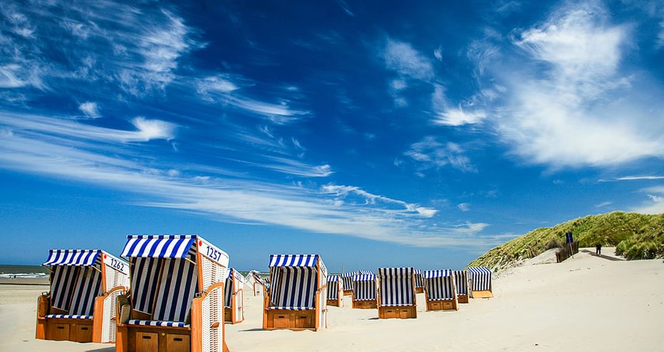 Reihe blau-weiß gestreifter Strandkörbe am Sandstrand mit Dünen unter blauem Himmel auf den Ostfriesischen Inseln.