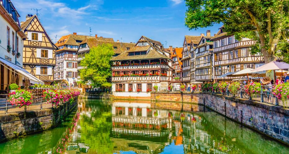 Traditionelle Fachwerkhäuser spiegeln sich in einem Kanal in Straßburg. Bunte Blumen säumen die Steinmauer unter blauem Himmel.