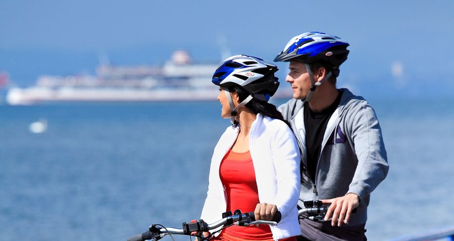 Zwei Radfahrer mit Helmen stehen mit ihren Rädern am Bodensee und blicken auf eine Fähre auf dem blauen Wasser mit Bergen im Hintergrund.