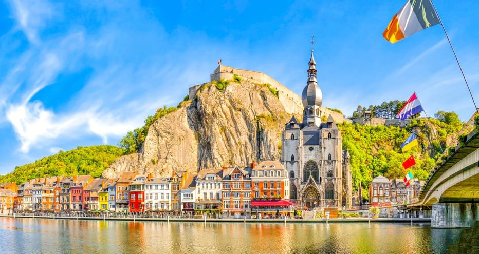 Stadtansicht von Dinant mit bunten Häusern an der Maas, gotischer Kirche und Zitadelle auf Felsenklippe unter blauem Himmel mit belgischer Flagge.
