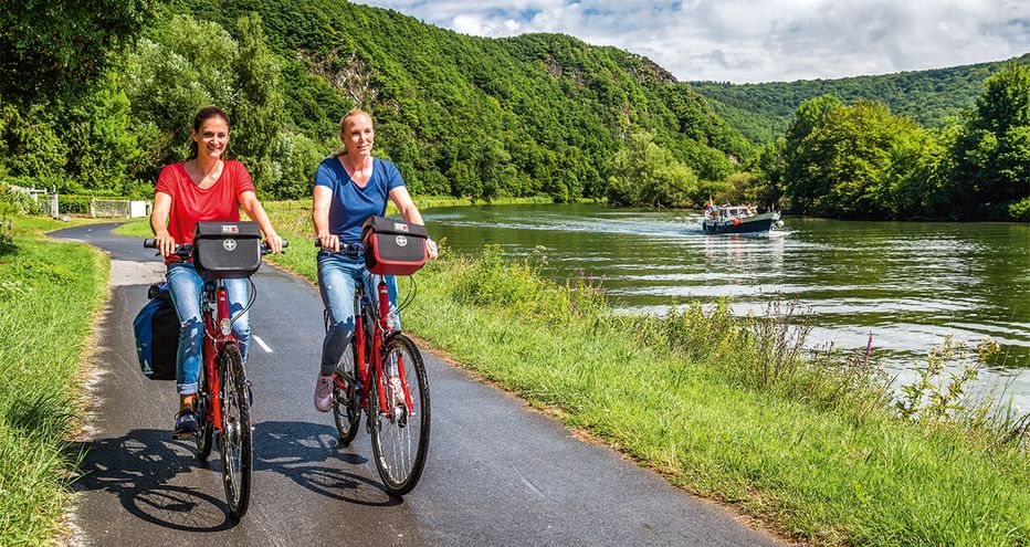 Zwei Frauen radeln auf asphaltiertem Weg entlang eines Flusses in Luxemburg mit bewaldeten Hügeln und Ausflugsboot im Hintergrund.