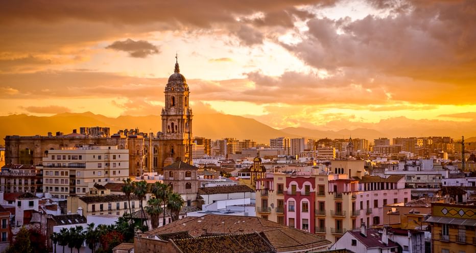 Sonnenuntergang über Málaga in Andalusien mit Kirchturm über weißen Gebäuden. Orangefarbener und goldener Himmel mit Bergen im Hintergrund.