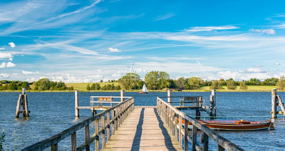 Holzsteg, der in die Schlei führt, mit festgemachten Booten. Blaues Wasser, grünes Ufer mit Bäumen und Windrädern unter hellem Himmel.