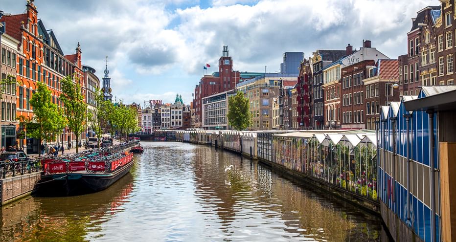Amsterdamer Gracht gesäumt von bunten historischen Gebäuden unter bewölktem Himmel. Eine Brücke überquert das Wasser, Boote liegen am Ufer.