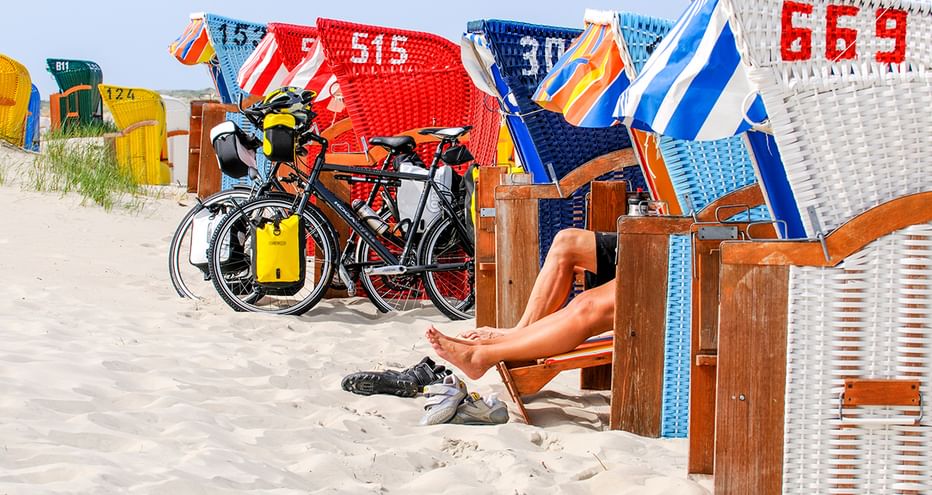Beine einer Person in Strandkorb am Sandstrand mit Tourenrad und gelben Packtaschen. Bunte Strandkörbe im Hintergrund.