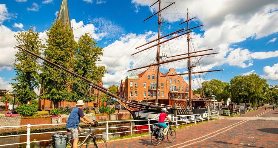 Zwei Radfahrer auf einem Backsteinweg neben einem historischen Segelschiff in Papenburg. Kirchturm und orangefarbene Backsteingebäude unter blauem Himmel.