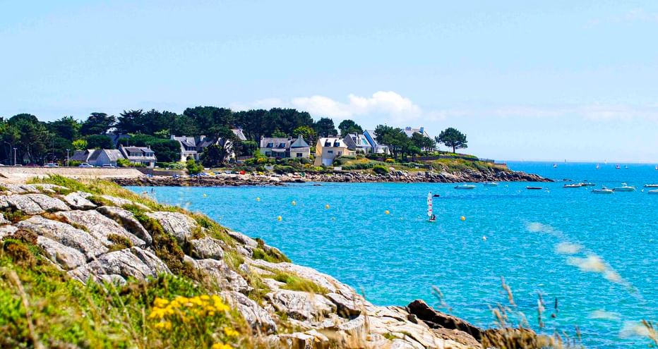 Felsige Küste mit gelben Wildblumen bei Saint-Colomban. Blaues Meer mit Booten und Bojen, weiße Häuser und Bäume am gegenüberliegenden Ufer.