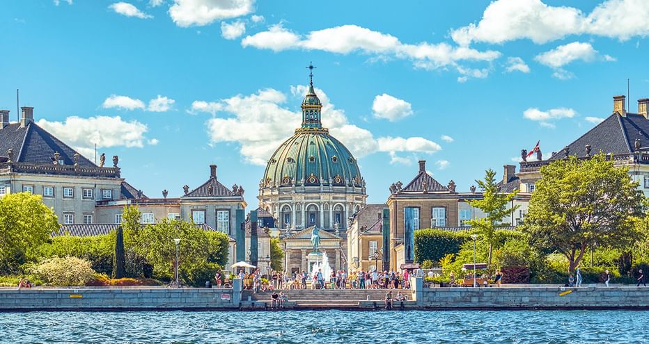 Blick über das Wasser zur Frederikskirche mit ihrer markanten blaugrünen Kupferkuppel, umgeben von historischen Gebäuden und Bäumen unter blauem Himmel.