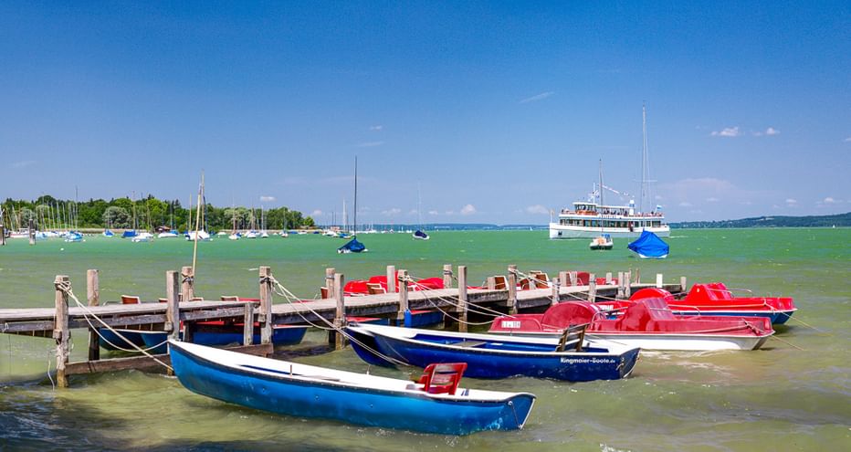 Blaue und rote Tretboote an einem Holzsteg an einem bayerischen See. Grünes Wasser, Segelboote und ein weißes Ausflugsschiff im Hintergrund.