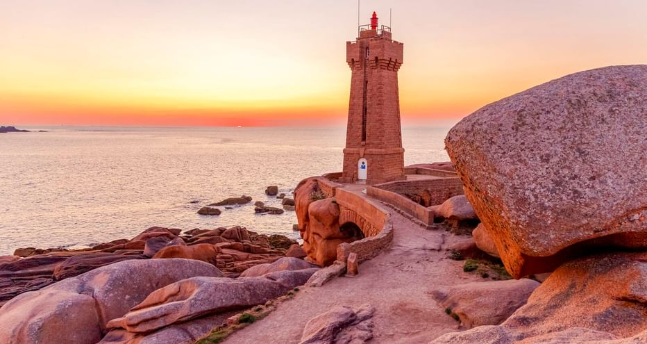 Rosa Granitleuchtturm von Ploumanac'h bei Perros-Guirec, umgeben von großen rosa Felsen bei Sonnenuntergang mit Meer und orangefarbenem Himmel.