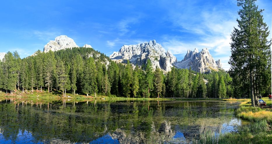 Klarer Bergsee spiegelt dramatische Dolomitengipfel und dichten Nadelwald unter blauem Himmel mit leichten Wolken.