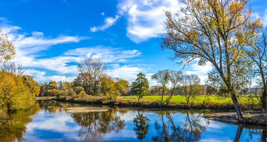 Calm river with perfect reflections of trees and blue sky in Bavaria. Green meadows and autumn trees line the riverbank along the Five Rivers Route.
