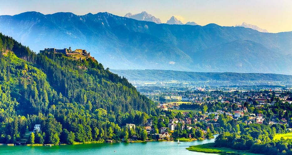 Türkisfarbener See mit bewaldeten Hügeln, Burg auf Hügel, Stadt im Tal und Bergkette unter blauem Himmel am Alpe Adria Radweg.