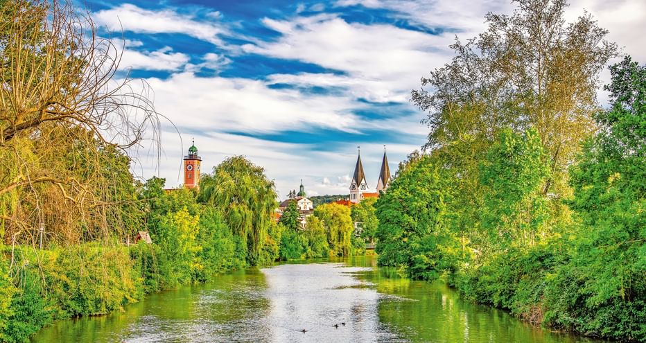 Blick auf die Altmühl in Eichstätt mit üppig grünen Bäumen am Ufer und historischen Kirchtürmen im Hintergrund unter blauem Himmel.