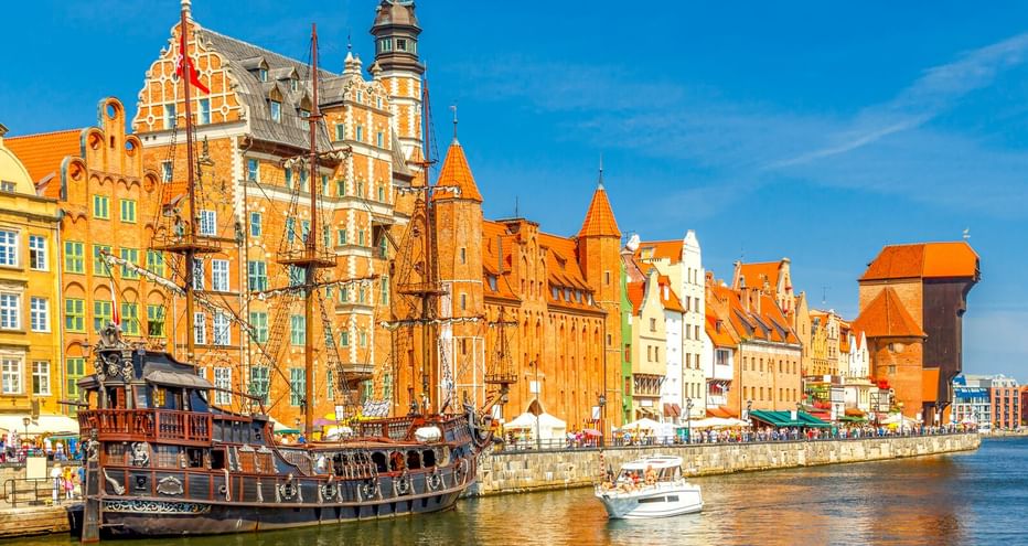 Bunte historische Gebäude entlang der Danziger Uferpromenade mit Segelschiff-Nachbau und mittelalterlichem Kranturm unter blauem Himmel.