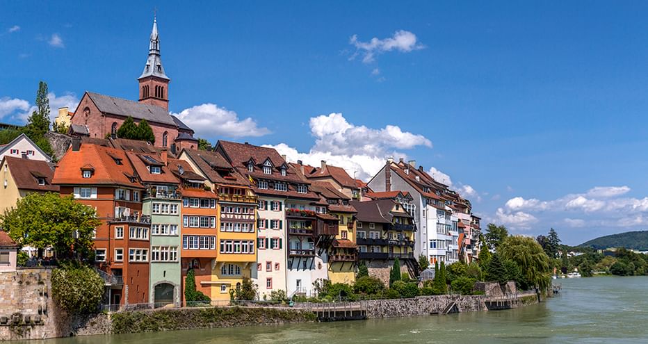 Historische bunte Häuser am Rhein mit Kirchturm auf dem Hügel. Blauer Himmel mit weißen Wolken über der Flussstadt.