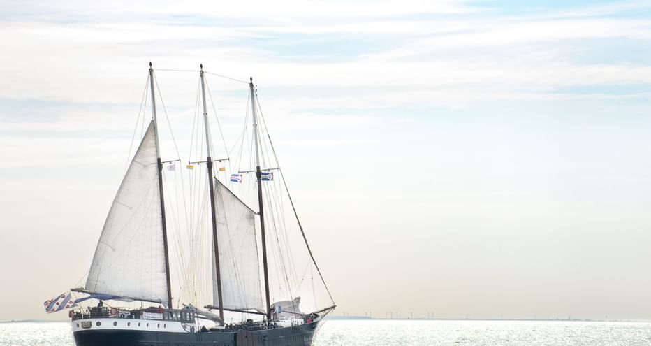 Dreimaster-Segelschiff mit weißen Segeln auf ruhigen Gewässern des Mare fan Fryslân unter bewölktem Himmel in den Niederlanden.
