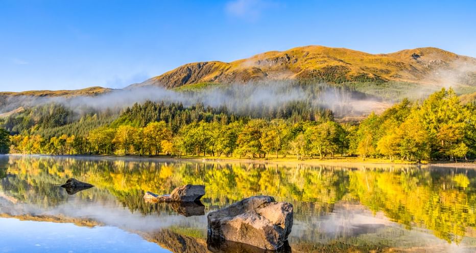 Ruhiger See in Highland Perthshire spiegelt Felsen, Bäume und goldene Hügel. Morgennebel schwebt über der bewaldeten Uferlinie unter blauem Himmel.