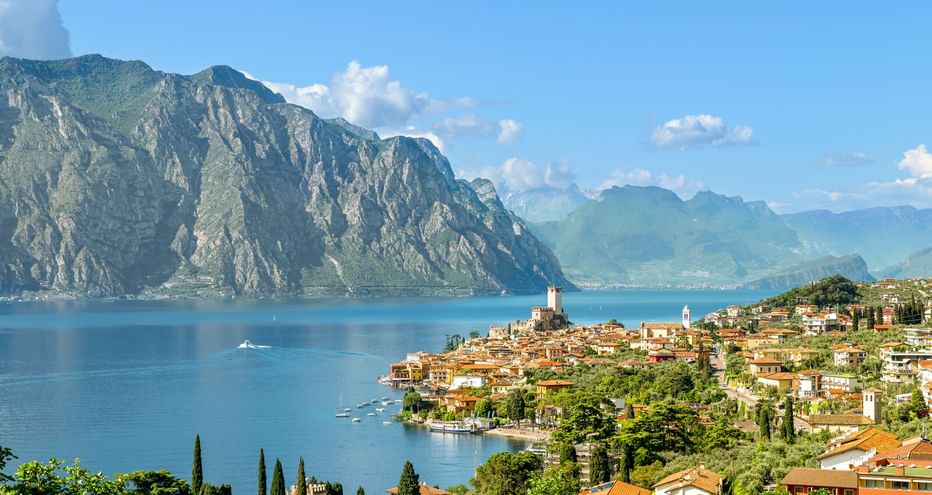 Panoramablick auf den Gardasee mit einer Hanglage-Stadt mit Terrakotta-Dächern, Kirchturm und dramatischen Bergen am blauen Wasser.