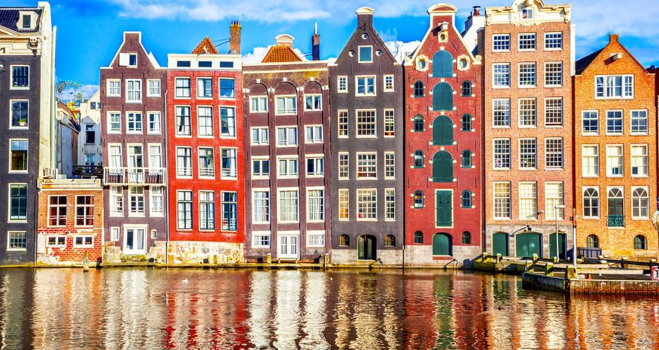 Reihe traditioneller niederländischer Grachtenhäuser in verschiedenen Farben wie Rot, Braun und Orange, die sich im Wasser der Amsterdamer Gracht spiegeln.
