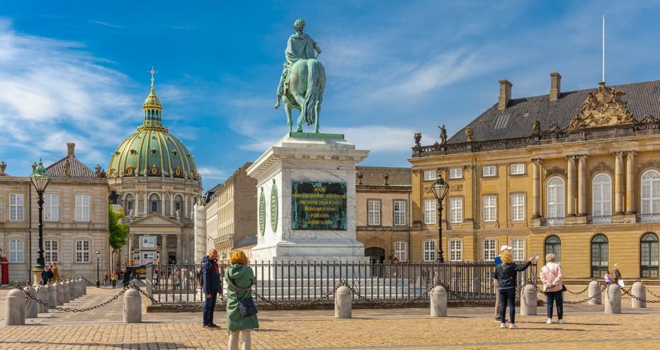 Amalienborg Schlossplatz in Kopenhagen mit Reiterstatue, gelben Palastgebäuden und Kirchenkuppel unter blauem Himmel mit Touristen.