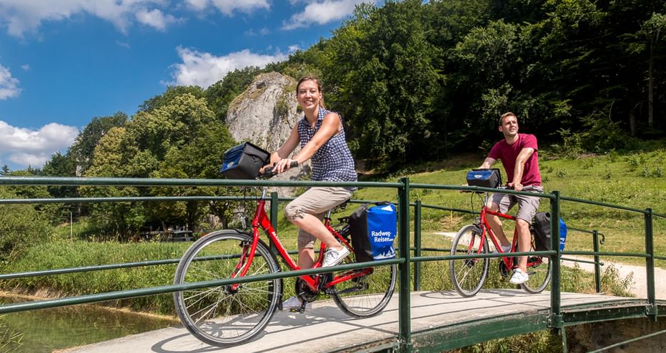 Zwei Radfahrer mit bepackten Rädern überqueren eine Metallbrücke über einen Kanal. Grüne Hügel und Wald im Hintergrund unter blauem Himmel.