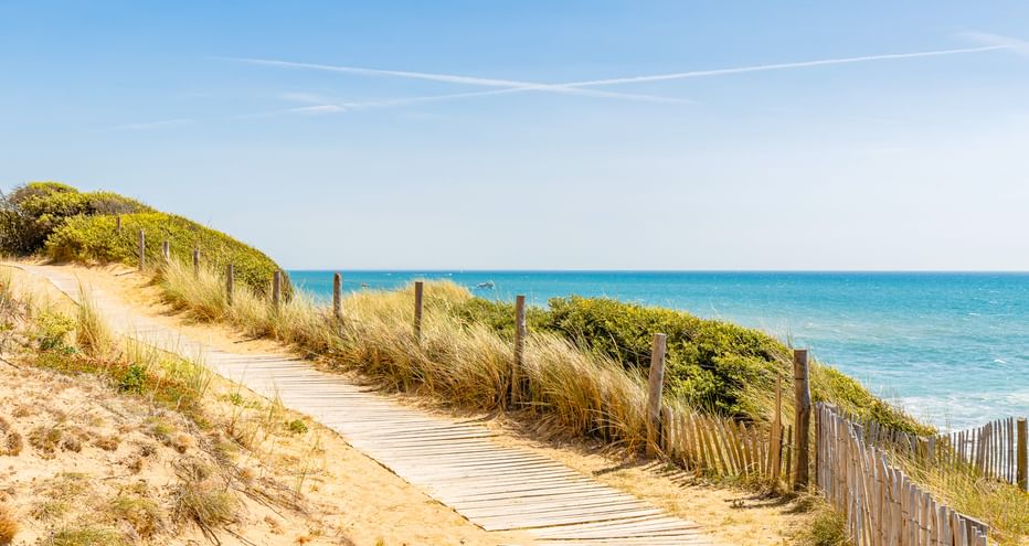 Holzsteg schlängelt sich durch Sanddünen mit Strandgras an der Pointe du Payre bei Jard-sur-Mer. Holzzaun säumt den Weg, türkisfarbenes Meer sichtbar.