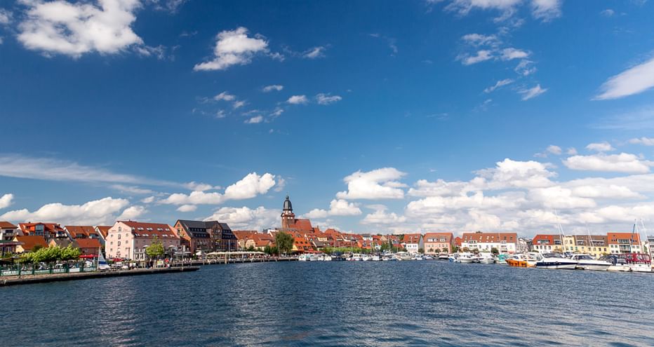 Panoramablick auf eine historische Stadt am Müritz-Ufer mit rotgedeckten Gebäuden, Kirchturm und Booten am Ufer unter blauem Himmel.