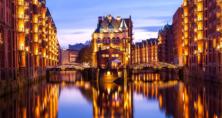 Historische rote Backsteingebäude bei Dämmerung beleuchtet, spiegeln sich im Kanalwasser. Brücken verbinden die Gebäude der Speicherstadt.