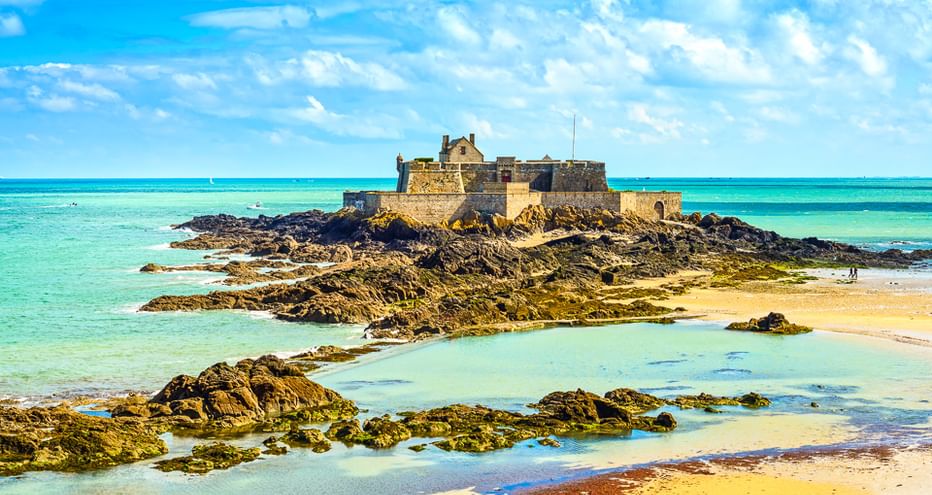 Steinfestung auf felsigem Vorsprung, umgeben von türkisfarbenem Wasser und freiliegenden Felsen bei Ebbe in der Nord-Bretagne unter blauem Himmel.
