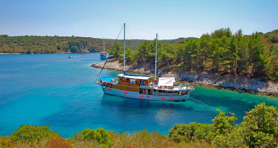 Holzmotoryacht MS Mirabela vor Anker in türkisfarbener Bucht, umgeben von grünem Pinienwald an der kroatischen Küste unter blauem Himmel.