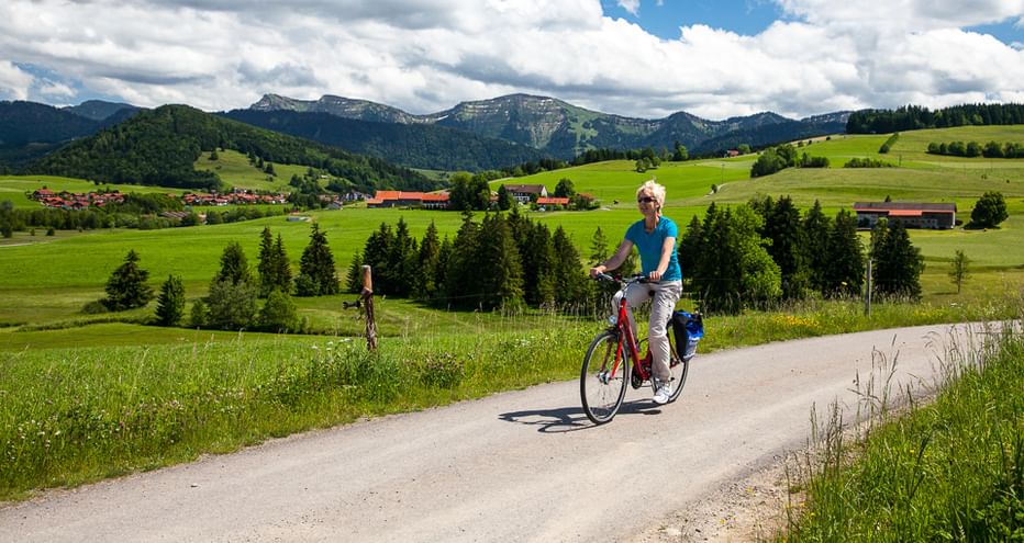 Frau radelt auf asphaltierter Landstraße durch grüne Allgäuer Landschaft mit Bergen, Wiesen, Wäldern und Dorf im Hintergrund unter bewölktem Himmel.