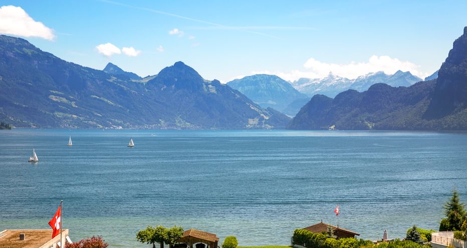 Vierwaldstättersee mit Segelbooten auf türkisfarbenem Wasser, umgeben von Bergen. Schweizer Flagge im Vordergrund mit grüner Wiese und Häusern am Ufer.