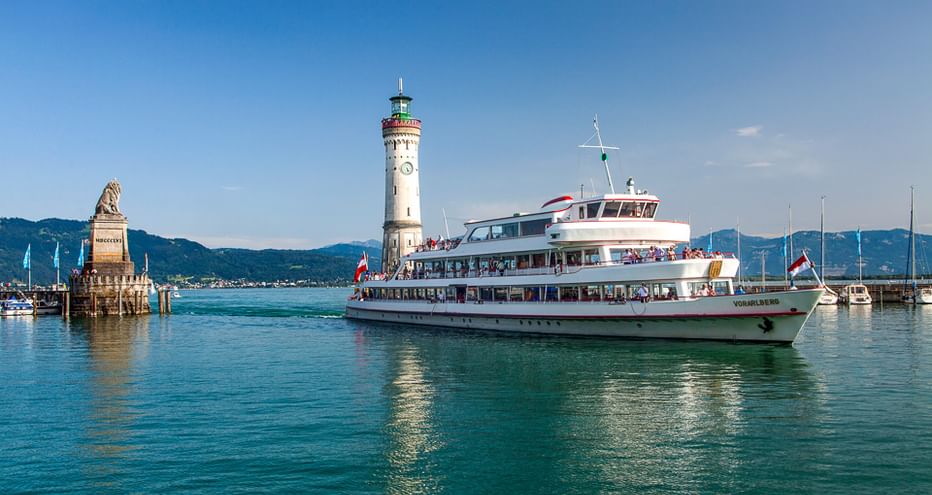 Weiße Personenfähre auf dem Bodensee passiert die Lindauer Hafeneinfahrt mit Leuchtturm und Löwenstatue. Berge im Hintergrund.