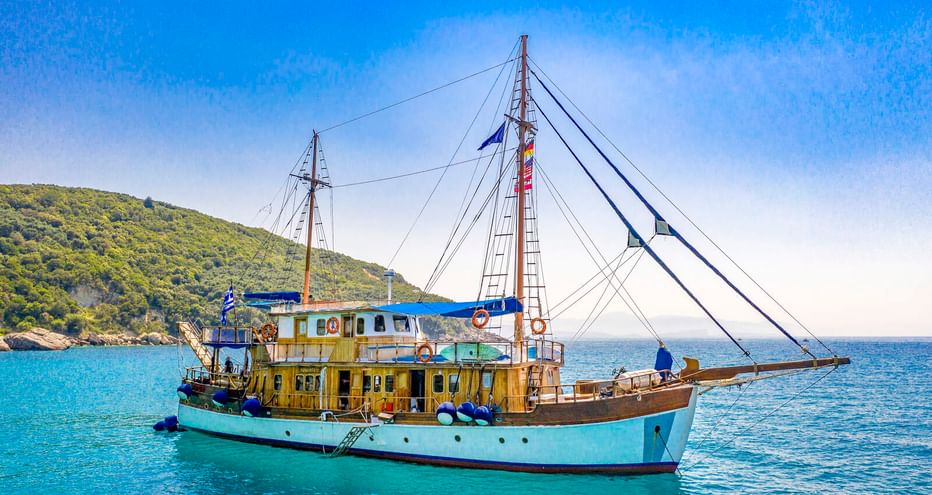 MS Panagiota, ein traditionelles Segelschiff mit türkisfarbenem Rumpf und Holzdeck, vor Anker in ruhigem blauen Wasser bei Parga mit grünen Hügeln.
