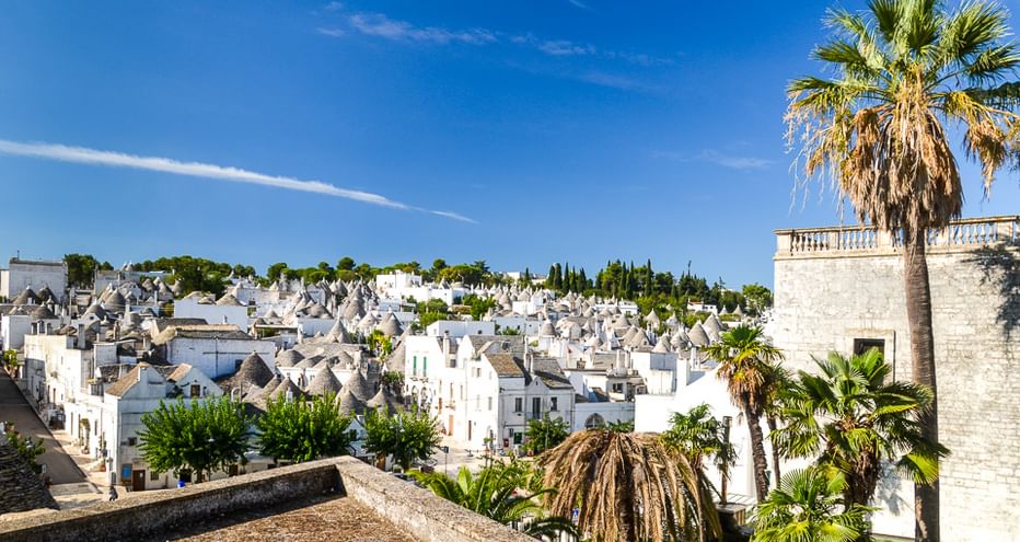 Blick auf traditionelle weiße Trulli-Häuser mit Kegeldächern in Apulien, Italien. Palmen im Vordergrund, blauer Himmel über dem historischen Dorf.