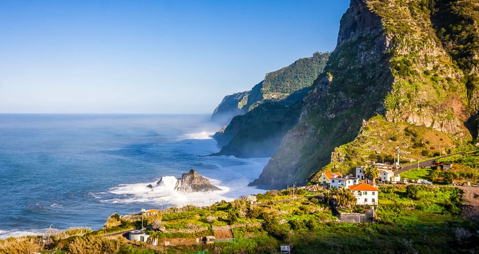 Weiße Häuser auf grünen Terrassenhängen unter steilen Klippen auf Madeira. Wellen brechen an Felsen entlang der blauen Atlantikküste.