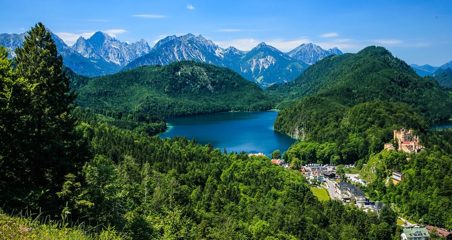 Luftaufnahme des Alpsees umgeben von bewaldeten Hügeln, Dorf Hohenschwangau mit Schloss und schneebedeckten Alpengipfeln unter blauem Himmel.