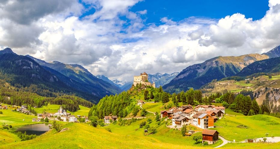 Schloss Tarasp auf einem Hügel, umgeben von grünen Wiesen und Dorfhäusern, mit Bergen und blauem Himmel im Hintergrund.