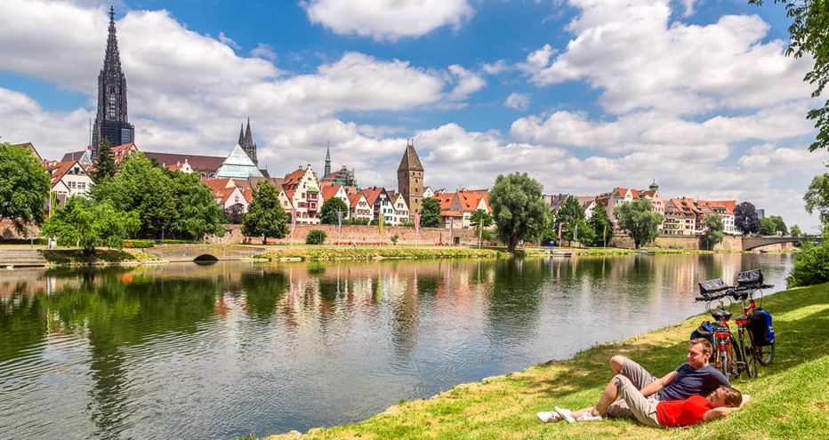 Zwei Radfahrer rasten am grasbewachsenen Donauufer mit Fahrrädern in der Nähe. Historische Stadt mit Kirchtürmen und bunten Gebäuden spiegelt sich im Wasser.