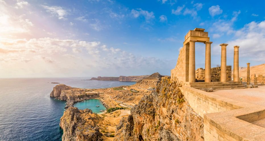 Antike griechische Säulen der Lindos Akropolis auf einer Klippe mit Blick auf das Mittelmeer und die felsige Küste mit kleiner Bucht.