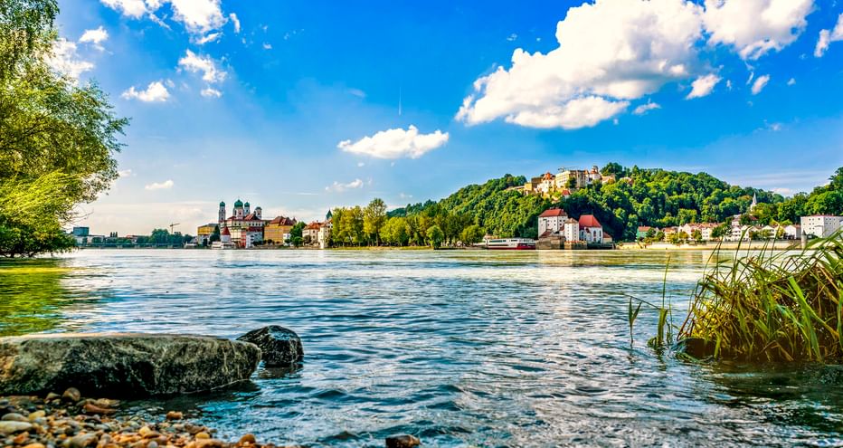 View across the Danube River in Passau with pebble shore, historic buildings and cathedral on the left, and a hilltop fortress under blue sky.