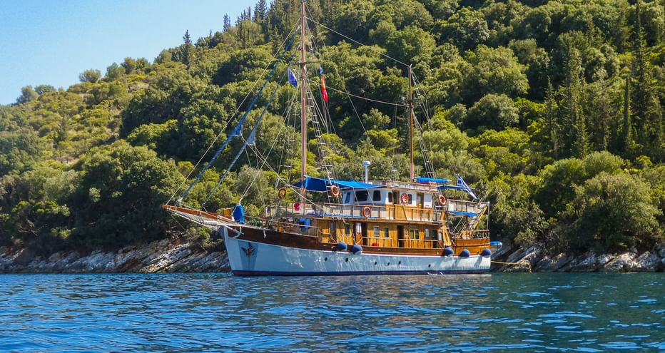 MS Panagiota, ein traditionelles Holzsegelschiff mit weiß-blauem Rumpf, vor Anker in ruhigem blauen Wasser nahe der bewaldeten griechischen Küste.