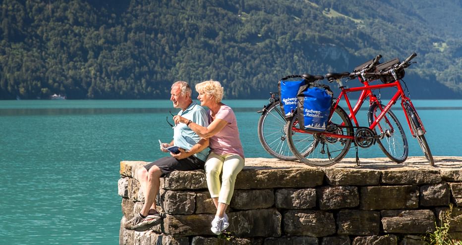 Zwei Radfahrer sitzen auf einer Steinmauer neben türkisfarbenem Schweizer See mit Tourenrädern. Bewaldete Berge im Hintergrund.