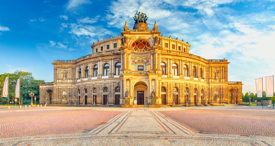 The Semperoper opera house in Dresden with its ornate Neo-Renaissance facade in golden sunlight, featuring arched windows and sculptures on top.