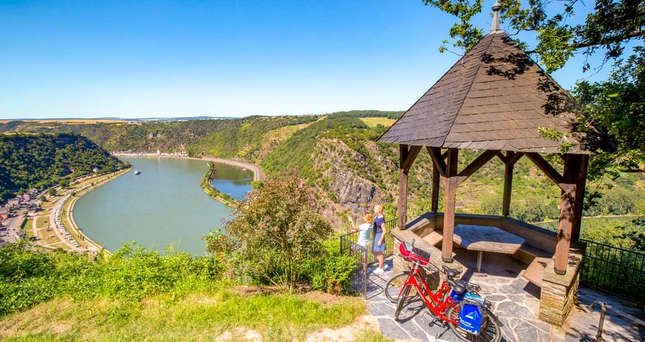 Wooden gazebo at Maria Ruh viewpoint with red bicycles, overlooking the Rhine River valley. Green hills and a curved river are visible below.