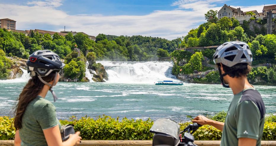 Zwei Radfahrer mit Helmen betrachten den Rheinfall von einer Aussichtsplattform. Ein Ausflugsboot fährt im türkisfarbenen Wasser unterhalb des breiten Wasserfalls.