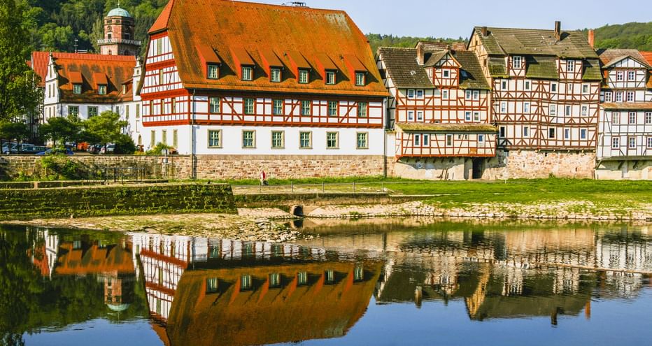 Traditionelle Fachwerkhäuser mit orangen Dächern entlang der Fulda in Rotenburg, die sich im ruhigen Wasser spiegeln, mit grünen Hügeln dahinter.