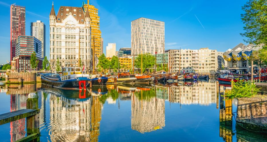 Rotterdamer Hafen mit Booten im ruhigen Wasser, das moderne Wolkenkratzer und das historische Witte Huis unter blauem Himmel spiegelt.