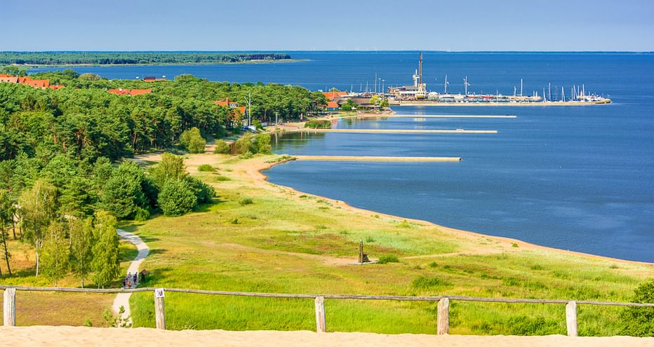Panoramablick auf die litauische Küste mit grünen Wiesen, Sandstrand, blauem Buchwasser und Marina mit Segelbooten in der Ferne.